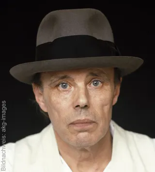 A portrait photograph of the German artist Joseph Beuys, wearing his characteristic grey felt hat, against a dark background.