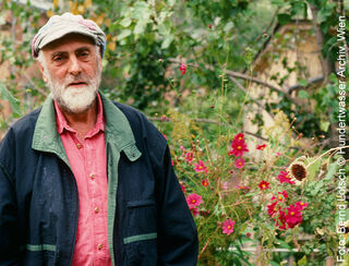 The Austrian artist and painter Friedensreich Hundertwasser, wearing a cap and sporting a beard, standing in a garden full of pink flowers.