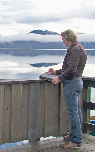 The painter Ansgar Skiba is standing on a wooden jetty by a lake, sketching a landscape.