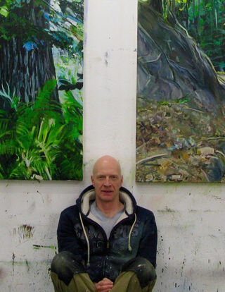 The German artist and painter Burkhard Kern, bald, sitting in his studio in front of two of his large-scale, naturalistic forest and landscape paintings.